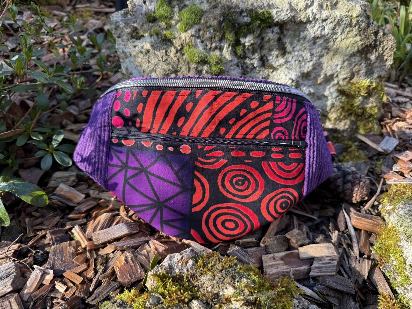 Banane Charly en velours violet et wax rouge à spirales, posée sur la pierre moussue