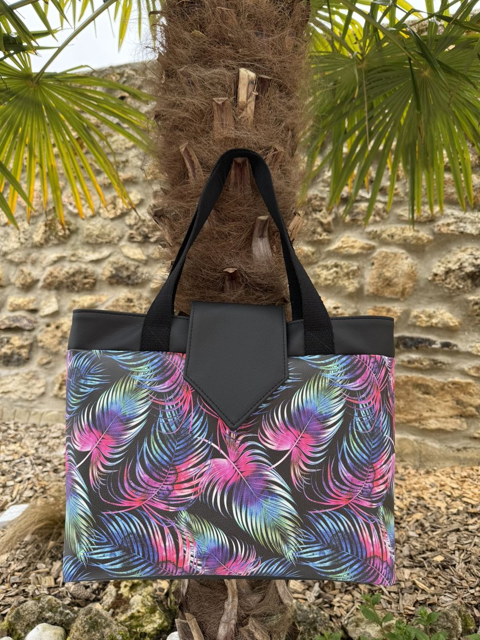 Sac Madison à palmes roses et bleues, anses noires, suspendu à un palmier